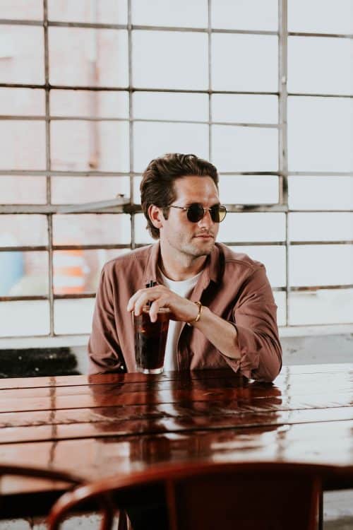 Homme en t-shirt blanc, veste marron et lunettes de soleil en train de boire un verre à une table