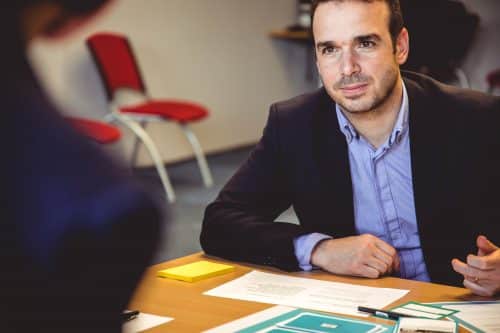 homme assis devant un bureau avec des papiers portant une chemise bleue et une veste de costume sombre